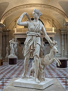 A marble statue of the Greek goddess Diana, depicted in classical style. She stands gracefully, with one hand raised as if holding a bow, while her other hand rests gently on the back of a deer at her side. The deer gazes upward, embodying the connection between the goddess and the natural world. Diana, the goddess of the hunt, is shown wearing a flowing tunic and a quiver of arrows on her back. The figure is detailed with delicate features, and her expression is serene yet focused, embodying both strength and tranquility. The statue captures a moment of harmony between the goddess and the animal, symbolizing her role as protector of wildlife