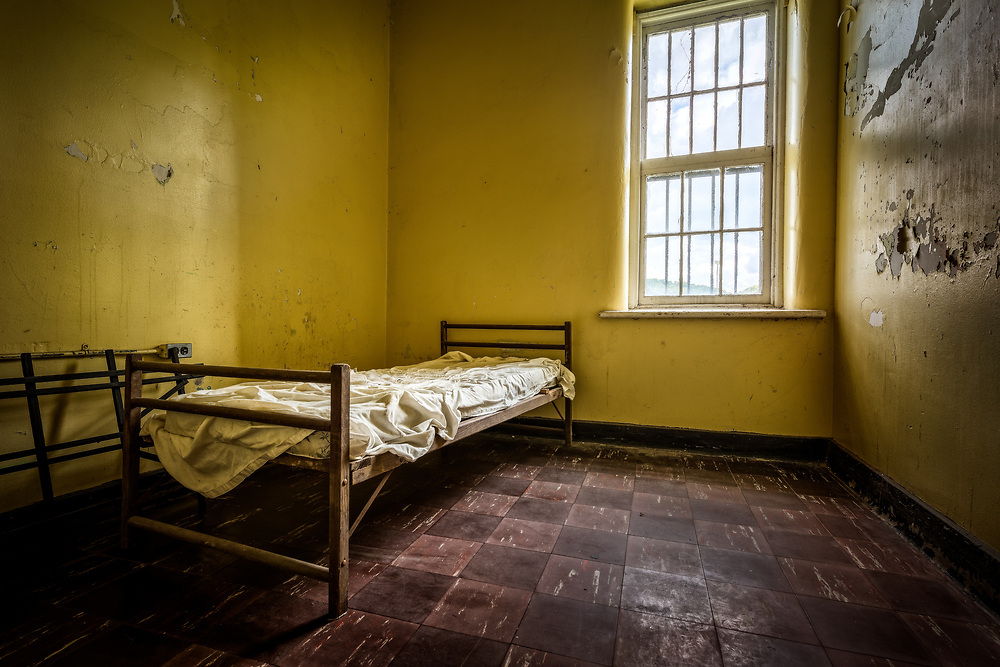 Small room in a mental institution. Has brown flooring, one barred window, yellow paint on the walls that is peeling and one small wooden bed frame with a small mattress on top. 