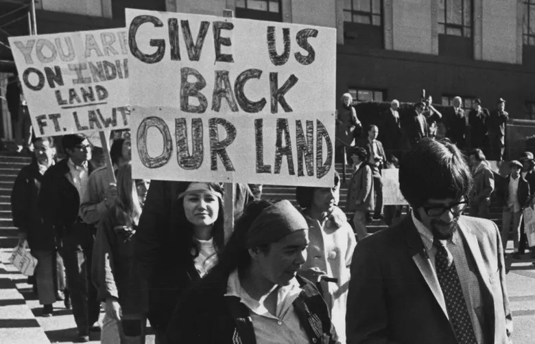 A black-and-white photo of a Native American protest, showing an individual holding a sign that reads 'Give Us Back Our Land.'