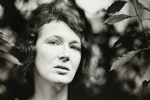 xAuthor Angela Carter standing in front of a backdrop of lush green leaves, smiling and looking towards the camera. She is dressed in a casual outfit, with her hair styled in a short, dark cut